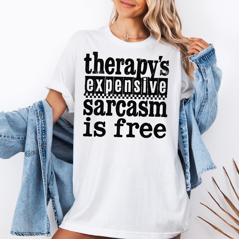 K&ouml;nnte beinhalten: Wei&szlig;es T-Shirt mit dem Text "therapy's expensive sarcasm is free" in schwarzer, fetter Schrift. Der Text ist in drei Zeilen angeordnet, mit einem Schachbrettmuster zwischen der zweiten und dritten Zeile. Das Shirt hat eine lockere Passform.
