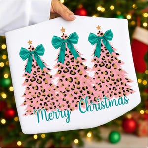 May include: White fabric with a pink leopard print Christmas tree design. Each tree has a teal bow and gold star. The words "Merry Christmas" are written in teal script.