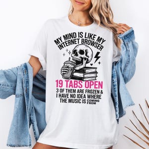 May include: White t-shirt with a graphic design featuring a skull holding a drink, a lightning bolt, and the text "MY MIND IS LIKE MY INTERNET BROWSER 19 TABS OPEN". The shirt also has the text "3 OF THEM ARE FROZEN & I HAVE NO IDEA WHERE THE MUSIC IS COMING FROM".