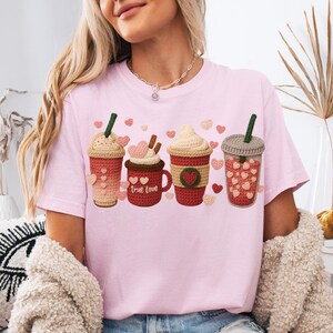 May include: A light pink t-shirt featuring four crochet coffee cup designs with heart accents. The cups are in shades of brown, red, and cream, with the words "True Love" on one cup. The shirt has a crew neck and short sleeves.