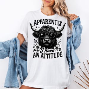 May include: White t-shirt featuring a black and white illustration of a Highland cow wearing sunglasses. The text "Apparently I have an attitude" is printed above and below the cow image. The shirt has short sleeves.