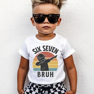 May include: A white t-shirt with rolled-up sleeves featuring the text "SIX SEVEN BRUH" above a silhouette of a person dabbing in front of a sunset design. The child is wearing black sunglasses and checkered shorts.