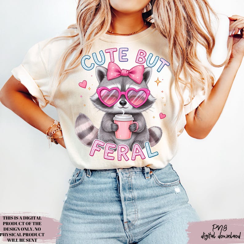 May include: Cream-coloured t-shirt with a cartoon raccoon wearing heart-shaped sunglasses and a pink bow. The raccoon holds a pink cup and the text reads "CUTE BUT FERAL". The design includes pink hearts and stars.