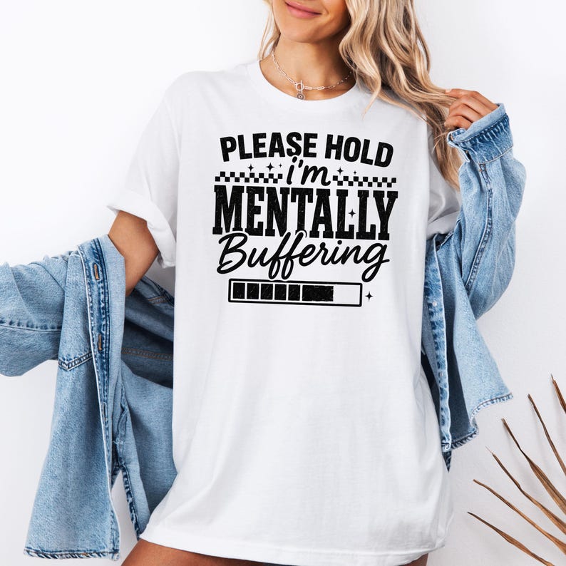 May include: White t-shirt with the text "PLEASE HOLD I'M MENTALLY BUFFERING" in black. The design includes a loading bar graphic. The shirt is a casual, unisex style.
