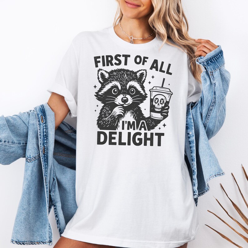 K&ouml;nnte beinhalten: Wei&szlig;es T-Shirt mit einer schwarz-wei&szlig;en Waschb&auml;r-Illustration, die eine Kaffeetasse mit einem Totenkopf-Design h&auml;lt. Der Text "FIRST OF ALL I'M A DELIGHT" ist &uuml;ber und unter dem Waschb&auml;r-Bild aufgedruckt. Das T-Shirt wird von einer Person getragen.