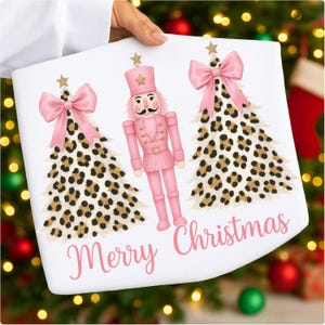 Può includere: Design a tema natalizio bianco con un soldato schiaccianoci rosa e due alberi di Natale con stampa leopardata. Gli alberi hanno fiocchi rosa e stelle dorate. Le parole "Merry Christmas" sono scritte in rosa.