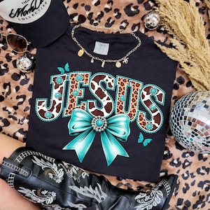 May include: Black t-shirt featuring the word "JESUS" in a cow print design, turquoise embellishments, and a turquoise bow. The shirt is styled with a gold necklace, sunglasses, and a black hat.