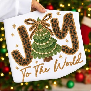 May include: White Christmas-themed pillow with the words "JOY To The World" embroidered in gold and leopard print. A green Christmas tree with gold garland and a bow is in the center, surrounded by gold snowflakes.