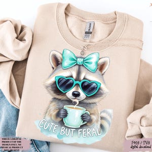 May include: Beige sweatshirt with a raccoon design. The raccoon wears heart-shaped sunglasses and a teal bow, holding a teal mug. The text "CUTE BUT FERAL" is below. A delicate necklace is visible near the collar.
