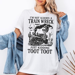 K&ouml;nnte beinhalten: Wei&szlig;es T-Shirt mit einem schwarzen Grafikdesign eines Skeletts, das einen Zug f&auml;hrt, mit dem Text "I'M NOT ALWAYS A TRAIN WRECK JUST KIDDING TOOT TOOT". Das Design beinhaltet einen Vollmond, Flederm&auml;use und Kakteen.