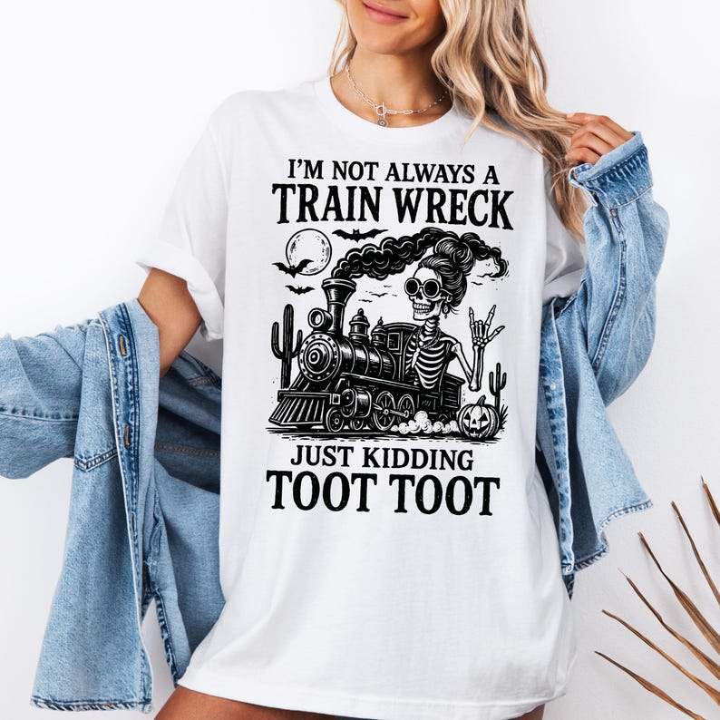K&ouml;nnte beinhalten: Wei&szlig;es T-Shirt mit einem schwarzen Grafikdesign eines Skeletts, das einen Zug f&auml;hrt, mit dem Text "I'M NOT ALWAYS A TRAIN WRECK JUST KIDDING TOOT TOOT". Das Design beinhaltet einen Vollmond, Flederm&auml;use und Kakteen.