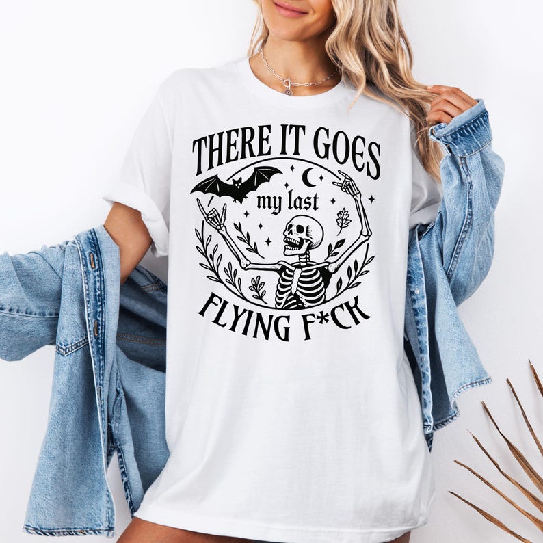 May include: White t-shirt with a black graphic design featuring a skeleton, bat, and the text "THERE IT GOES my last FLYING F*CK". The design includes stars and a crescent moon. The shirt has short sleeves.