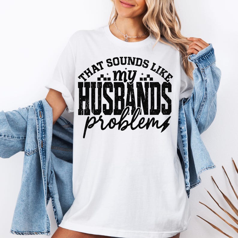 K&ouml;nnte beinhalten: Wei&szlig;es T-Shirt mit dem schwarzen Text "THAT SOUNDS LIKE my HUSBANDS problems". Das Shirt hat einen l&auml;ssigen Stil, der Text ist fett und gut lesbar. Eine Jeansjacke ist &uuml;ber den Schultern drapiert.