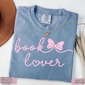 May include: A blue Comfort Colors t-shirt with the words "book lover" in pink script, featuring a bow and heart design. A gold necklace with a pendant is draped across the shirt. The shirt is a size small.