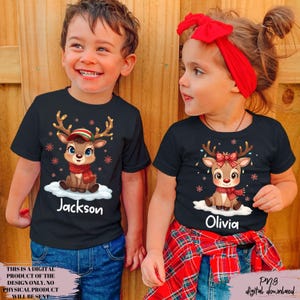 May include: Two children wearing black t-shirts with a cartoon reindeer design. The reindeer has antlers, a scarf, and a hat. The name "Jackson" is printed on one shirt, and "Olivia" on the other. The shirts are paired with blue jeans.