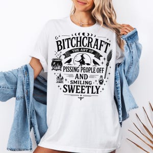 May include: White t-shirt with a black graphic design. The design includes the text "Bitchcraft The Art of Pissing People Off and Smiling Sweetly" with witch-themed illustrations such as a cauldron, witch on a broom, and crows.