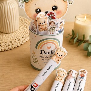 May include: A cylindrical container decorated with a rainbow and the German text Danke, dass du jetzt so viel schaffst. It holds several cut-out illustrations of bears and children. Four white tubes with the text du and various German phrases are held.