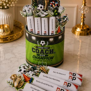 May include: A cylindrical container with a green soccer field design featuring the text "THANK YOU COACH, OUR CHAMPION ON AND OFF THE FIELD" and several soccer-themed toppers. Four white tubes with printed messages are included as accents.