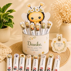 May include: A cylindrical container filled with white tubes featuring bee illustrations and German text. A large bee cutout sits atop the container. A separate bee-themed card reads "Danke für alles!" The container text reads "Danke, dass du jeden Tag Liebe säst." Additional tubes are arranged in front.