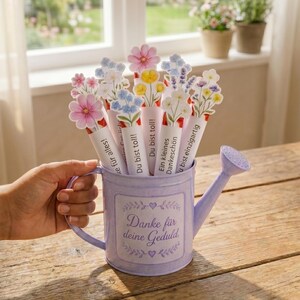 Puede incluir: Una regadera de metal de color morado claro con el texto Danke f&uuml;r deine Geduld y corazones decorativos. Dentro hay varios tubos de papel rematados con coloridas ilustraciones florales y frases en alem&aacute;n como Du bist toll y Ein kleines Dankesch&ouml;n.