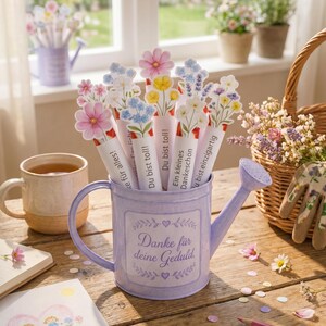 Puede incluir: Una regadera de metal de color morado claro con el texto Danke f&uuml;r deine Geduld, llena de varios recortes de flores de papel. Cada recorte muestra una ilustraci&oacute;n floral y frases en alem&aacute;n como Du bist toll y Ein kleines Dankesch&ouml;n.