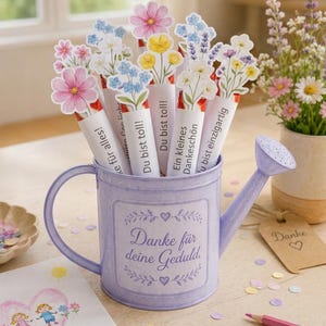 Puede incluir: Una regadera de metal de color morado claro llena de tubos rematados con flores de papel. Cada tubo presenta texto impreso en alem&aacute;n, incluyendo 'Danke f&uuml;r alles!', 'Du bist toll!', 'Ein kleines Dankesch&ouml;n' y 'Du bist einzigartig'. La regadera tiene la inscripci&oacute;n 'Danke f&uuml;r deine Geduld'.