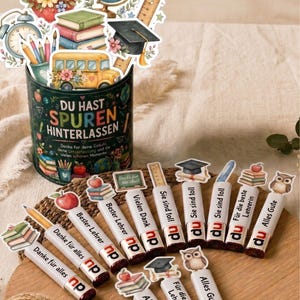 May include: A dark green cylindrical container with the text DU HAST SPUREN HINTERLASSEN, filled with school-themed cutouts like an apple, globe, books, and bus. Several chocolate bars with school-themed toppers and German text are arranged on a wooden board.
