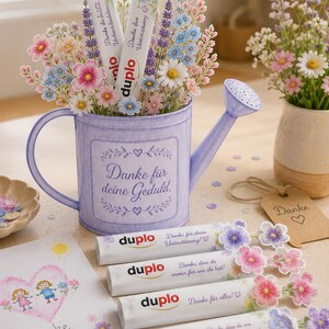 Puede incluir: Una regadera de metal de color morado claro llena de flores de papel y barritas de chocolate Duplo, con el texto 'Danke f&uuml;r deine Geduld'. Cuatro barritas Duplo adicionales con detalles florales y mensajes de agradecimiento en alem&aacute;n se encuentran delante, junto a una tarjeta con un dibujo de un coraz&oacute;n.