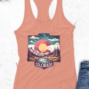 May include: A peach-colored tank top with a graphic of the Colorado state logo. The graphic features a stylized mountain landscape with a setting sun, birds, and the word "COLORADO" in a rectangular frame.