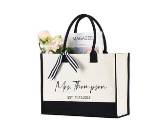 May include: A canvas tote bag with black handles and a black bottom panel. The bag features the text "Mrs. Thompson" and "EST. 11.15.2025" in a script font. A bouquet of flowers and a magazine are inside the bag.