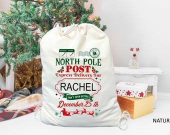May include: White Christmas gift bag with a red and green North Pole Post design. The bag has a personalized name, Rachel, and says "Express Delivery For". It also says "Don't Open Before December 25th".