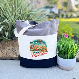 May include: A tri-color tote bag with a white center panel featuring a colorful Nebraska graphic. The bag has a gray top, a navy blue bottom, and white handles. The graphic includes the word "Nebraska" in red script.