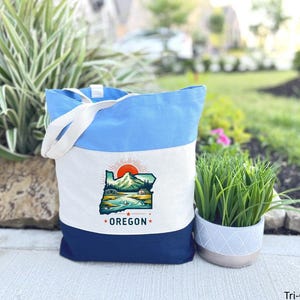 May include: A tri-color tote bag with a light blue top, white center, and navy blue bottom. The white section features a graphic of the Oregon state outline with a mountain, sun, and water scene. The word "OREGON" is below the graphic.