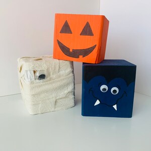 Könnte beinhalten: Drei Holzblöcke, die mit Halloween-Motiven bemalt sind. Der erste Block ist orange und bemalt, um wie eine Halloween-Laterne auszusehen. Der zweite Block ist weiß und bemalt, um wie eine Mumie auszusehen. Der dritte Block ist blau und bemalt, um wie ein Vampir auszusehen.