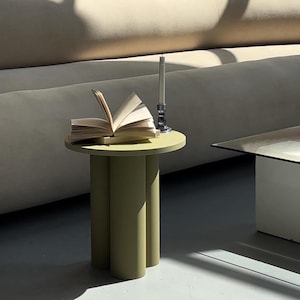May include: A small, round, olive-green side table with three cylindrical legs. An open book and a candle in a silver holder sit on top. The background features a beige sofa and a textured wall.