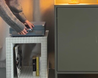 Puede incluir: Escena interior de estilo retro con una lámpara amarilla en forma de hongo y un jarrón naranja sobre un mueble gris. Un pequeño tocadiscos azul claro se encuentra sobre una mesa auxiliar de azulejos blancos con libros y revistas.