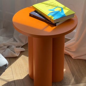 Puede incluir: Mesa auxiliar naranja con tapa redonda y tres patas cilíndricas. Dos libros apilados encima. Uno tiene una cubierta amarilla con gráficos azules y blancos, el otro una cubierta azul oscuro. La mesa mide unos 45 cm de diámetro.