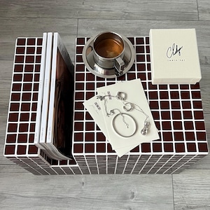 Pode incluir: Uma mesa de centro com um padrão de azulejos castanho e branco, com revistas, uma chávena de café prateada, uma caixa de joias com o texto "Chky" e joias. A mesa tem um porta-revistas embutido.