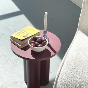 May include: A small, round, burgundy side table with three cylindrical legs. A stack of books with a yellow cover and the title "everything about love" sits on top, along with a white bowl of plums and a candle in a blue holder.