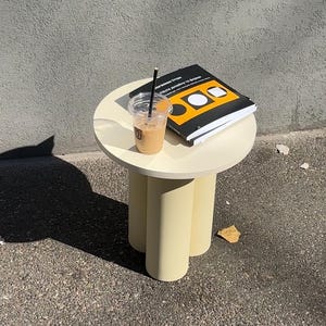 May include: A small, round, cream-colored table with three cylindrical legs. A clear plastic cup with iced coffee and a black straw sits on top, next to a book with a black cover and yellow and white geometric shapes. The table is outdoors.
