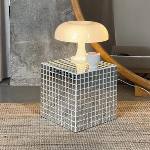 May include: A modern side table with a mirrored mosaic cube design. A white mushroom-shaped lamp and a small white cup sit on top. The table is placed on a woven rug.