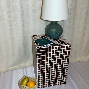 May include: A brown and white mosaic-tiled side table supports a teal lamp with a white shade and a small green book. A woven basket contains a pear and an orange. The table is a cube shape.