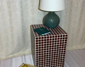 May include: A decorative side table with a mosaic tile pattern in brown and white. A teal lamp with a white lampshade sits on top, along with a small green book. A woven basket holds a pear and an orange.