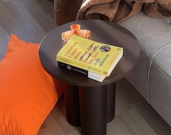 May include: A small, round, dark brown side table with three cylindrical legs. Two books and a hair clip rest on the table's surface. An orange cushion and a brown blanket are nearby.