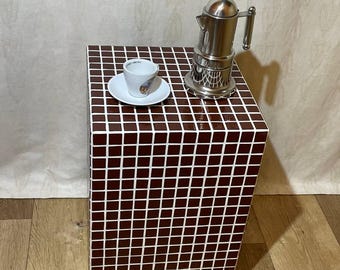 May include: A small, square side table with a brown and white tile pattern. A silver stovetop espresso maker and a white espresso cup and saucer are on top. The table is on a wooden floor.