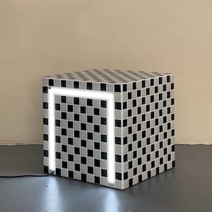 以下が含まれることがあります： 白黒の市松模様のキューブ型サイドテーブル。白い長方形のライトフレームが片側に組み込まれています。テーブルはコンセントに接続されています。