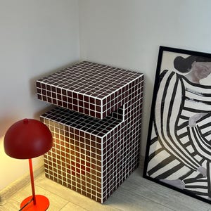 May include: A contemporary side table with a dark brown, tiled exterior and a stepped design. A small, red dome-shaped lamp with a matching base is positioned nearby. A framed black and white abstract artwork leans against the wall.