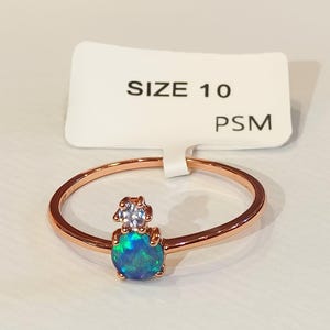 Op de afbeelding: Roségouden ring met een ronde, iriserende blauwgroene opaal en een klein, helder stenen accent. De ring is maat 10, zoals aangegeven op het aangehechte label. De ring ligt op een wit oppervlak.