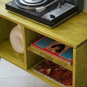 Puede incluir: Un mueble tocadiscos de madera verde lima con patas de horquilla negras. El mueble tiene un tocadiscos negro en la parte superior, un jarr&oacute;n blanco y estantes con discos de vinilo. El mueble es de madera y tiene un acabado texturizado.