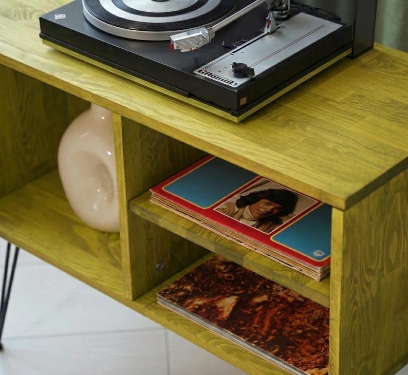 Puede incluir: Un mueble tocadiscos de madera verde lima con patas de horquilla negras. El mueble tiene un tocadiscos negro en la parte superior, un jarr&oacute;n blanco y estantes con discos de vinilo. El mueble es de madera y tiene un acabado texturizado.